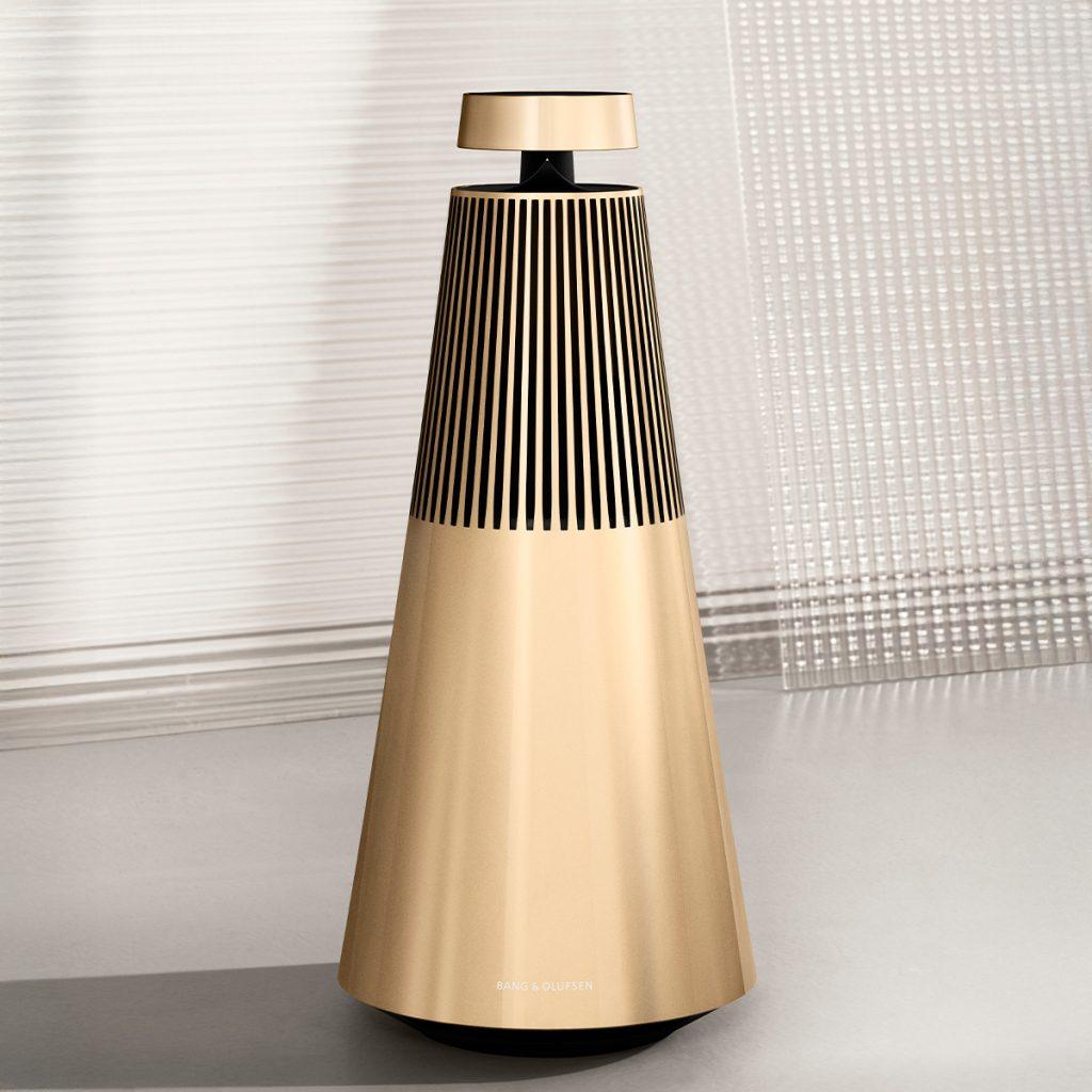 Bang & Olufsen Beosound 2 wireless speaker in gold finish, photographed against a modern textured background — luxury 360-degree audio system designed for premium interiors.