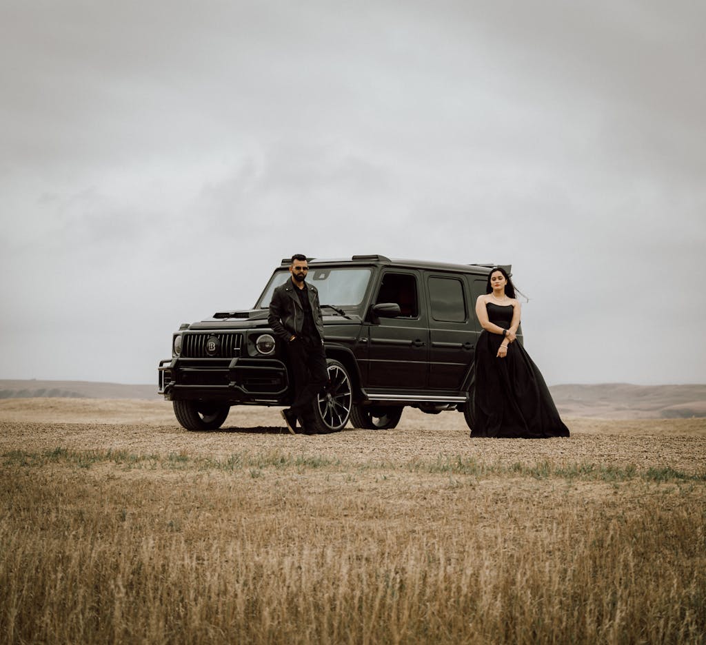 A fashionable man and woman standing with a black SUV in an open field on a cloudy day.