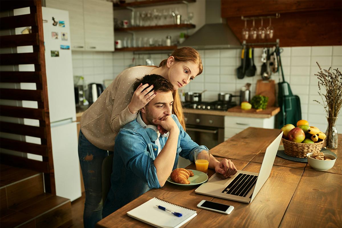 A young couple in their kitchen, reacting with concern while reviewing rising energy bills on a laptop — symbolizing the frustration many Australians feel about soaring electricity costs.