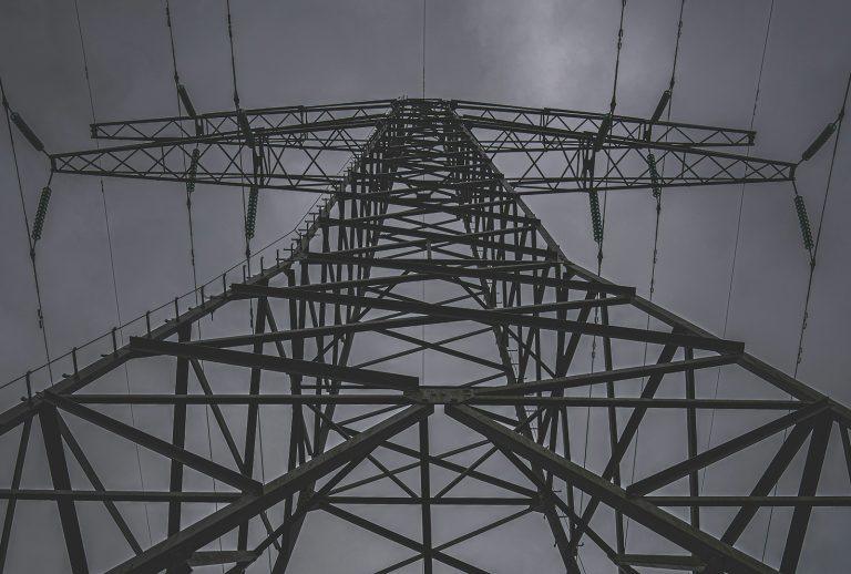 Looking up at a dark power transmission tower against stormy skies – a symbol of energy control and disillusionment.