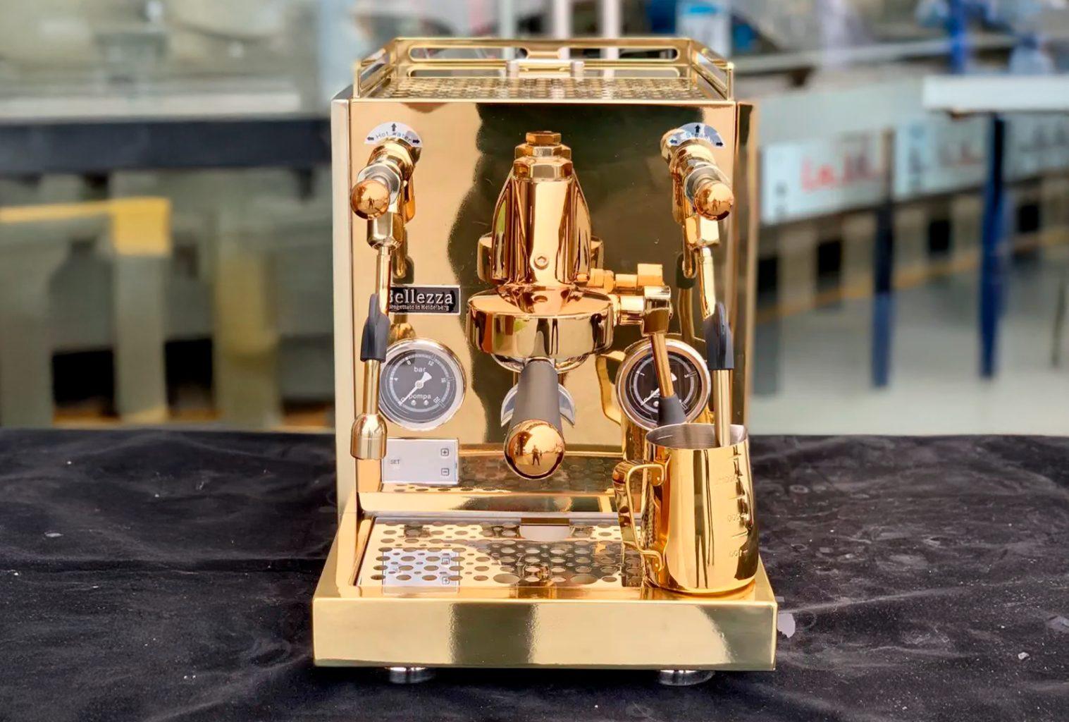 Bellezza Espresso Inizio espresso machine in polished 24K gold, front-facing on a black cloth backdrop, showcasing dual pressure gauges, polished metal wands, and a matching gold milk frothing jug.