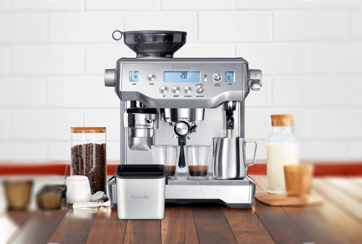 Stainless steel Breville Oracle espresso machine with twin shot glasses and milk jug, set against a clean white kitchen backdrop with wood benchtop, professional coffee setup for the home.