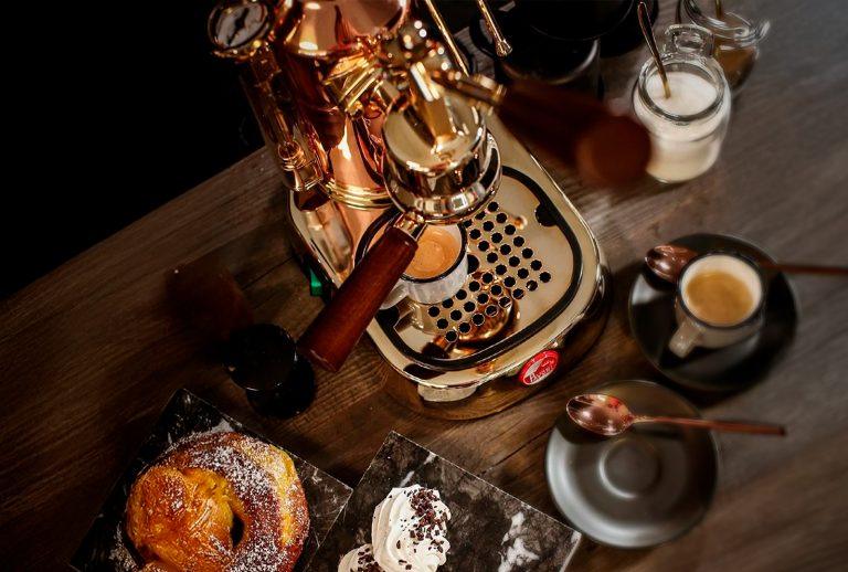 Top-down view of a copper and brass La Pavoni espresso machine pulling a shot, surrounded by fresh pastries, a ceramic espresso cup, and a wooden benchtop — a luxury home coffee setup.