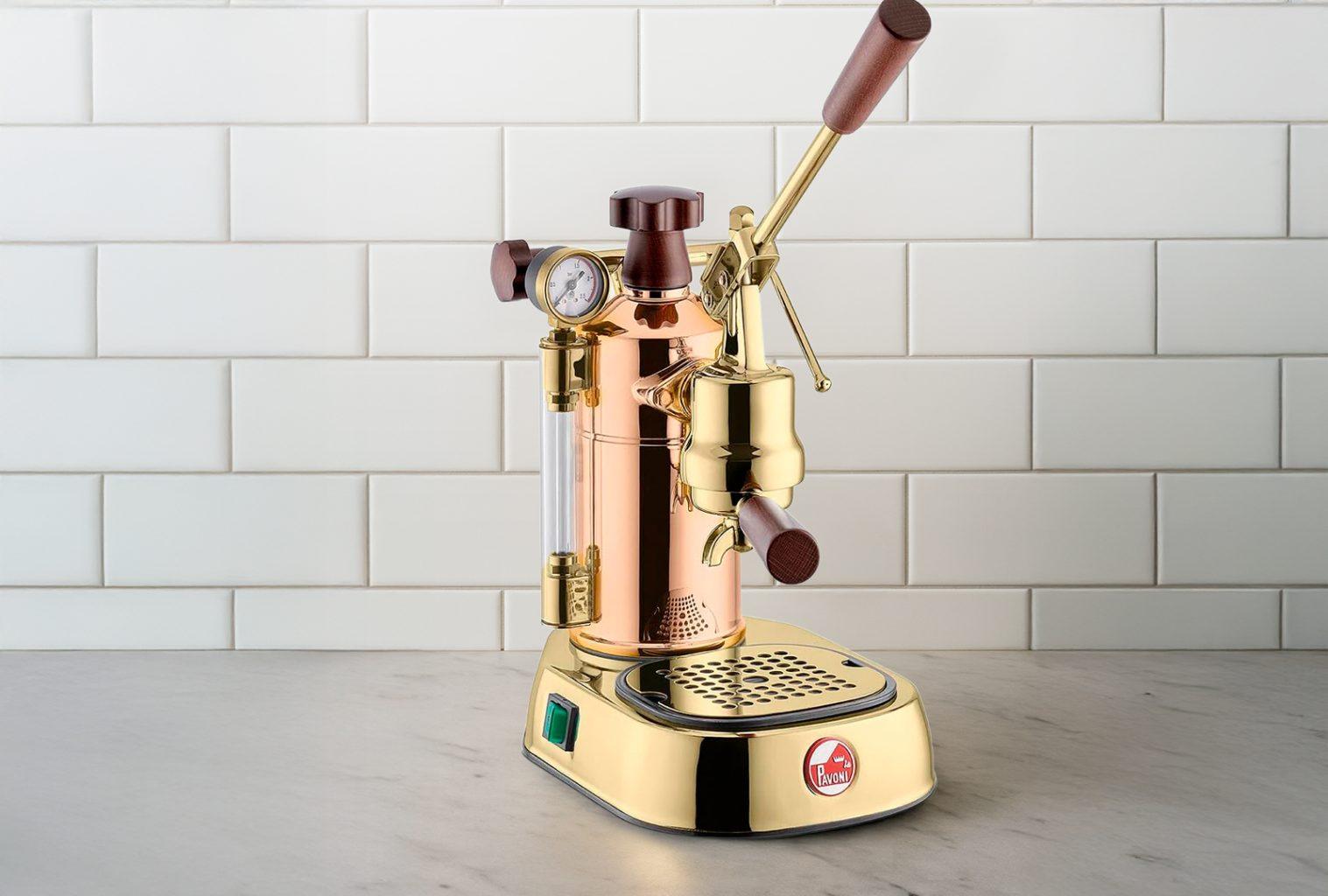 La Pavoni Professional lever espresso machine in polished copper and brass, positioned on a marble countertop against a white tiled backsplash — a minimalist, design-forward kitchen setup.