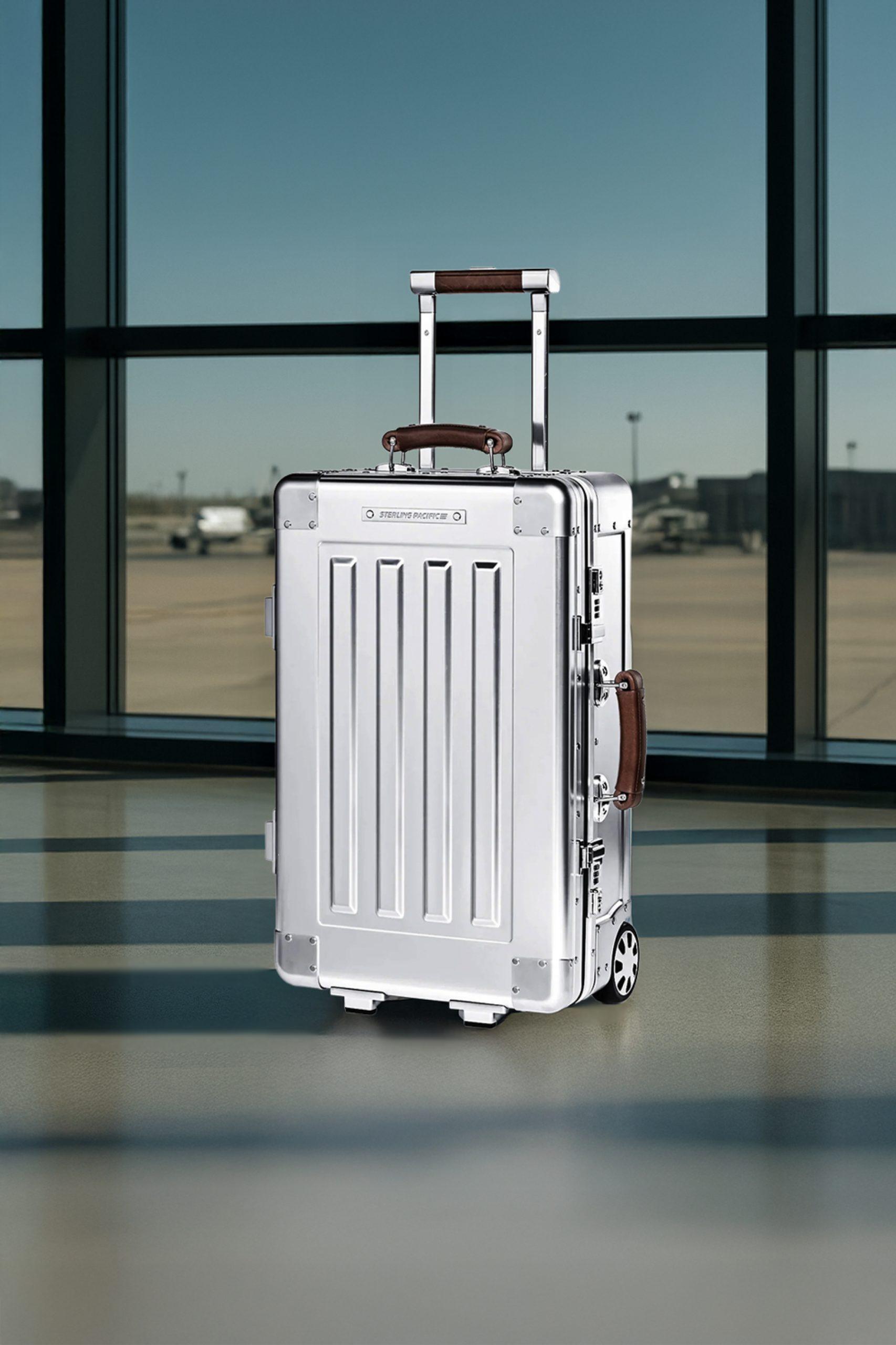 A brushed aluminum Sterling Pacific 35L cabin suitcase stands upright in an airport terminal with sunlight casting long shadows on the polished floor. Rich leather handles and industrial rivets highlight the craftsmanship of the luxury travel case.