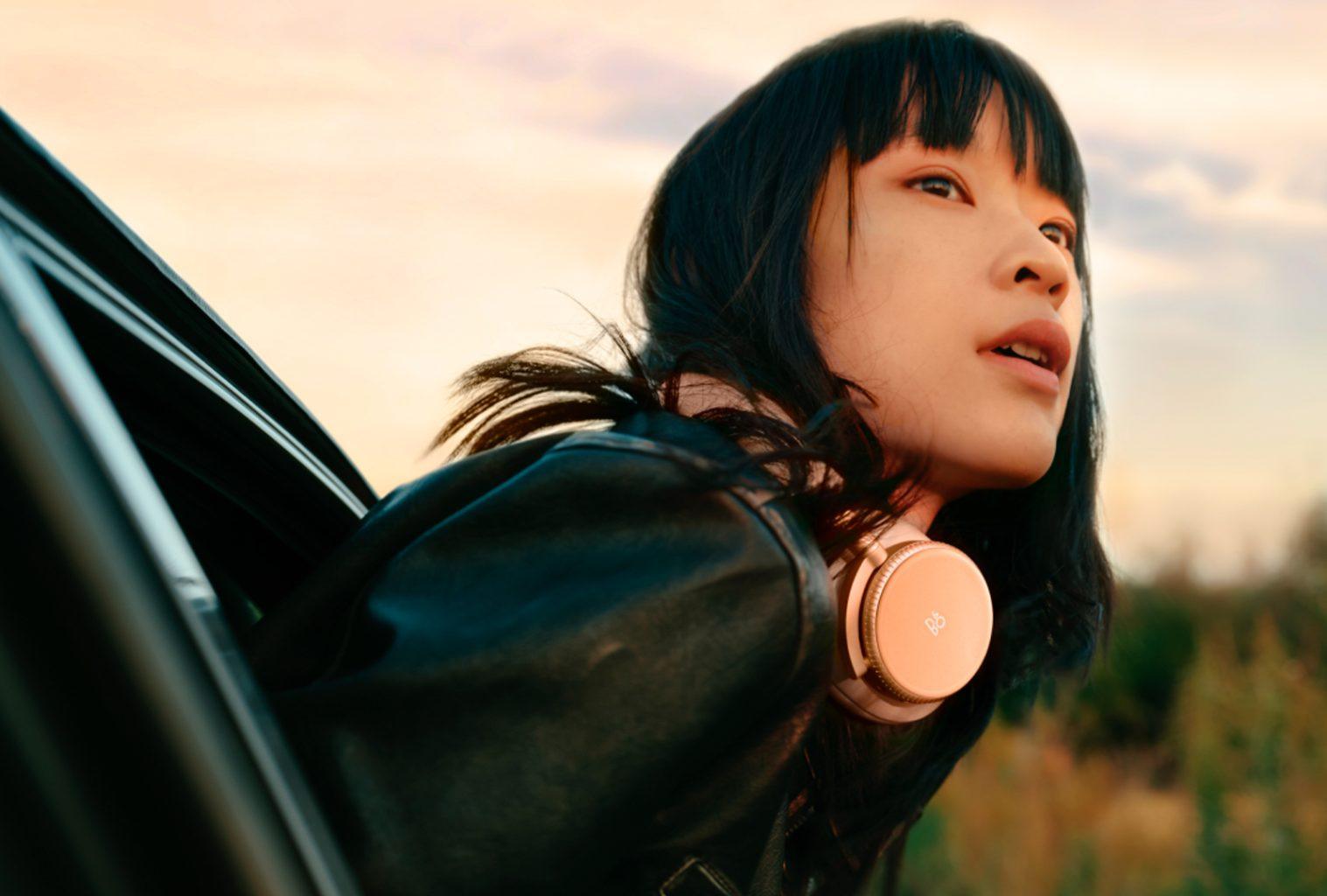 Woman wearing Bang & Olufsen Beoplay H100 headphones, leaning out of a car window at sunset, capturing a sense of freedom and immersive sound.