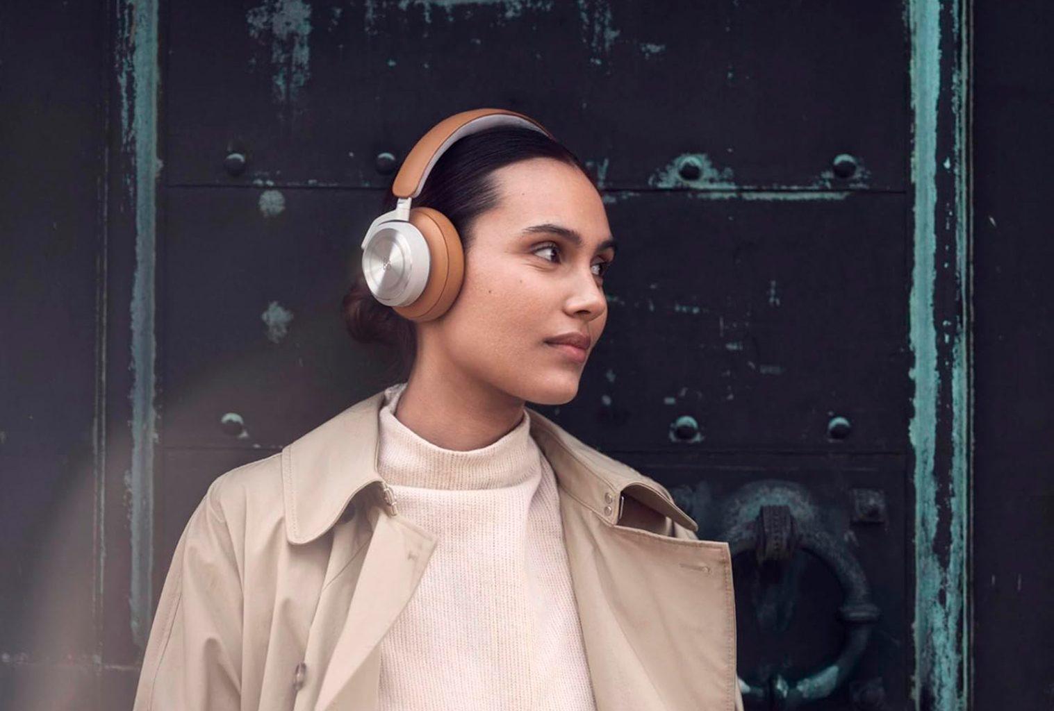 Woman wearing Bang & Olufsen Beoplay HX headphones in a neutral-toned outfit, standing against a dark architectural backdrop with teal accents.