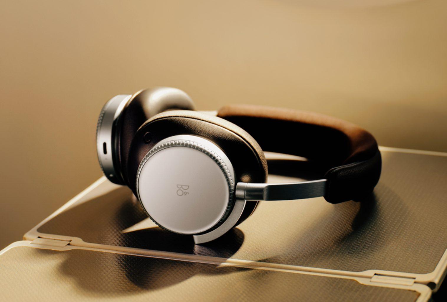 Bang & Olufsen Beoplay H100 wireless headphones resting on an airplane tray table, showcasing the minimalist build and precision-machined materials.