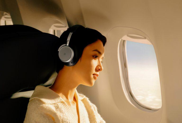 Woman wearing Bang & Olufsen Beoplay H100 headphones on an airplane, sitting by the window with soft sunlight highlighting the luxury design.