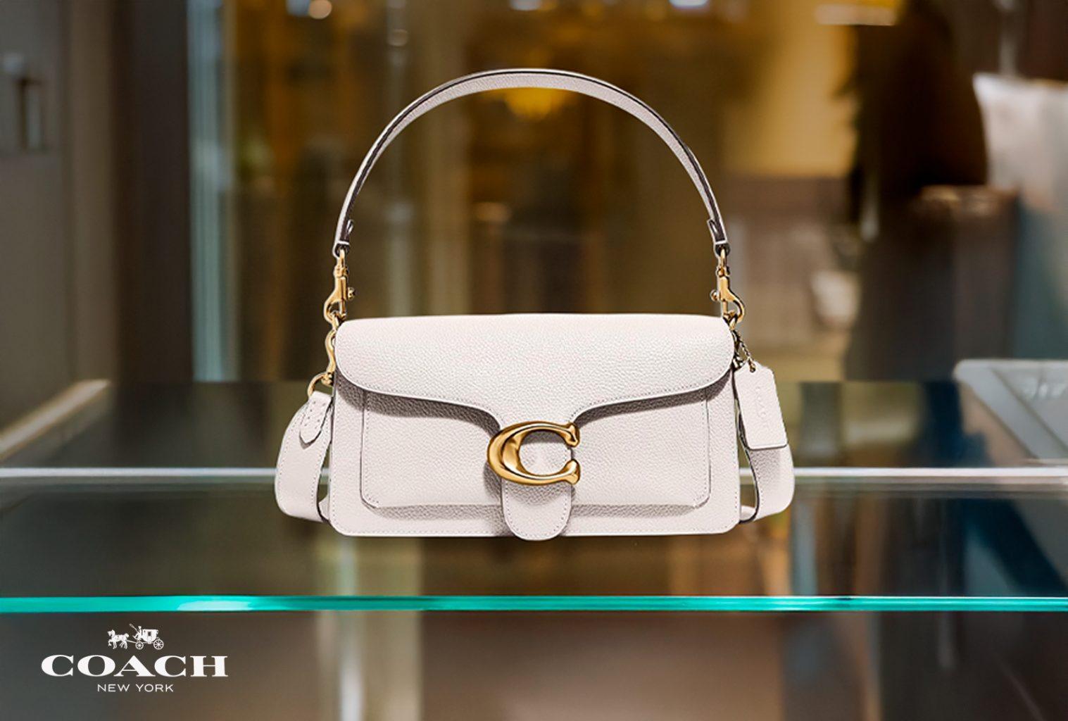Coach Tabby 26 shoulder bag in light taupe pebbled leather with gold 'C' logo, displayed on glass shelf.