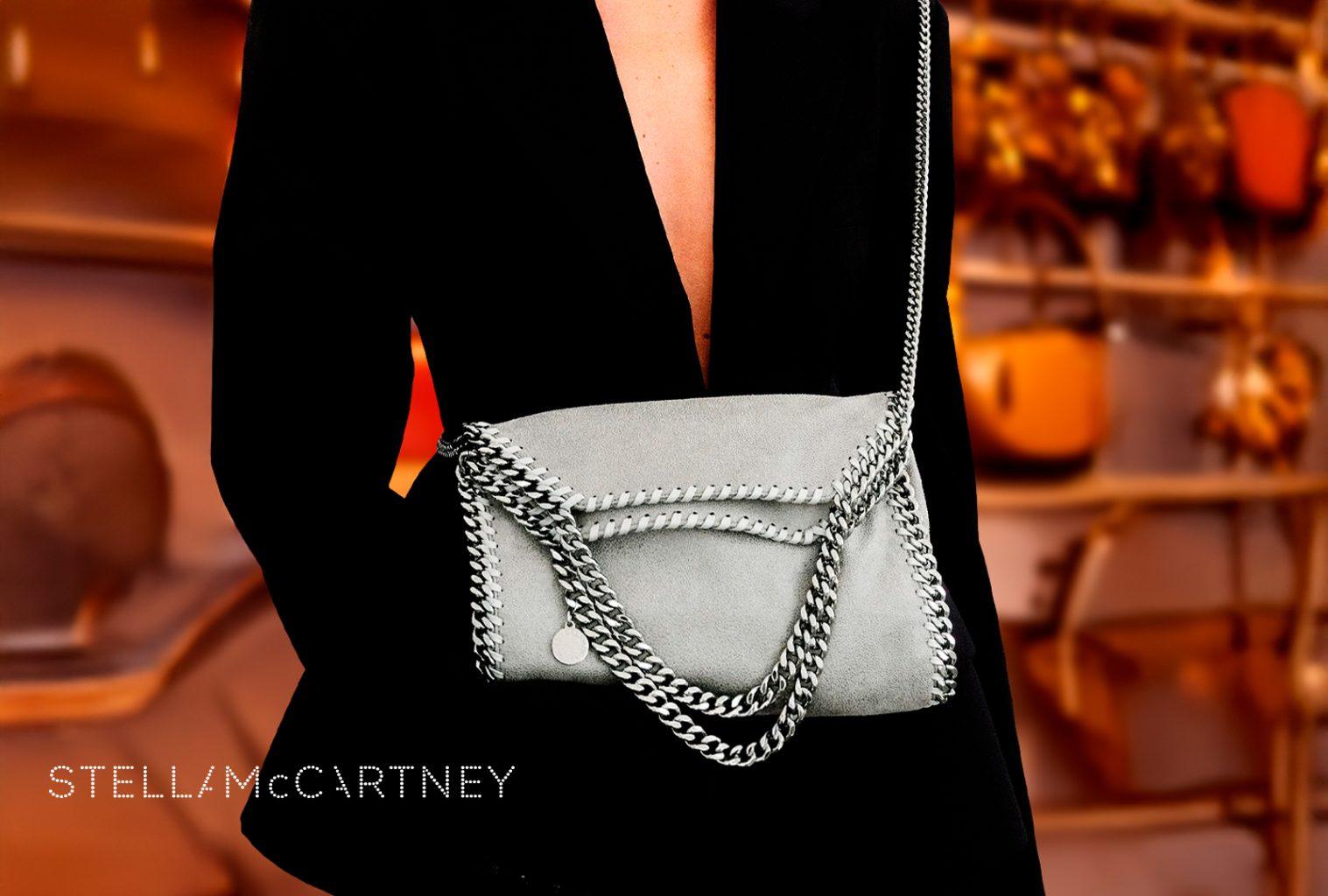 Model wearing black suit holding grey Stella McCartney Falabella bag with silver chain trim in boutique setting.