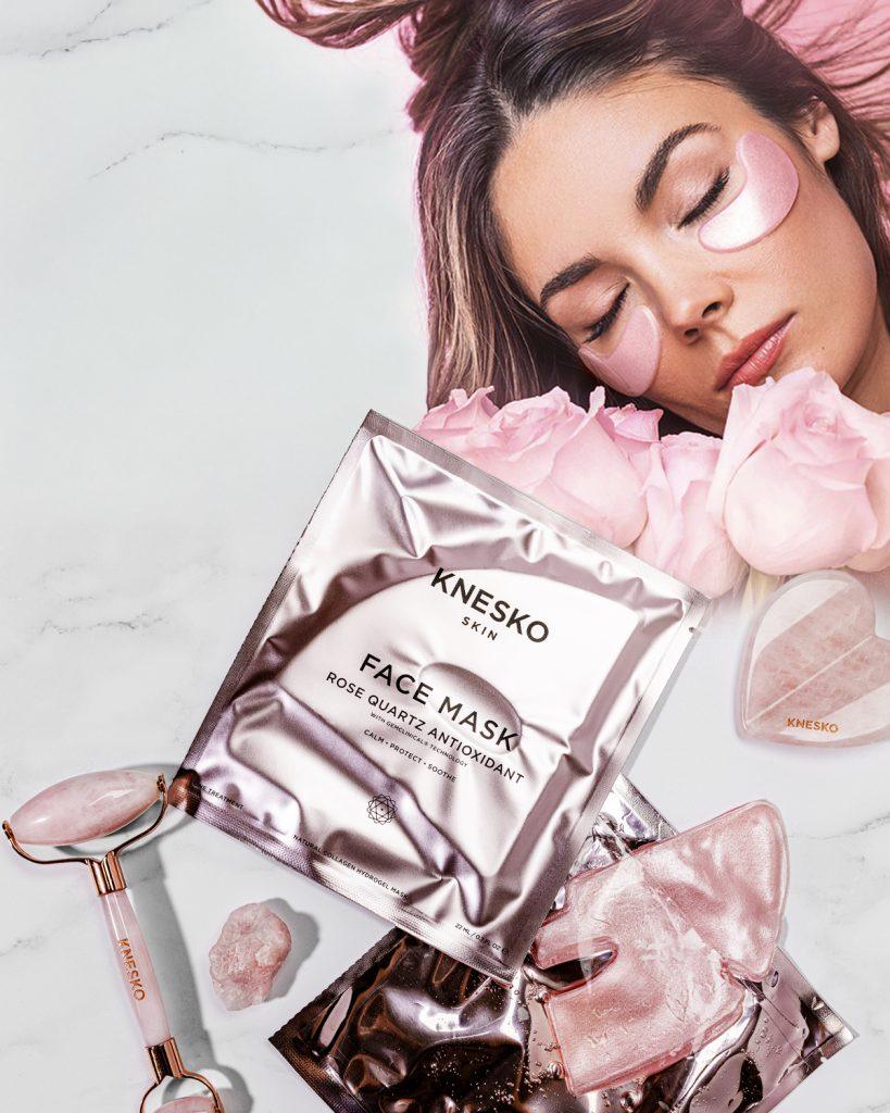 KNESKO Rose Quartz Collection featuring a woman relaxing with pink under-eye masks, surrounded by roses, face mask packets, a rose quartz roller, gua sha tool, and crystals — luxury skincare products designed to calm, protect, and soothe.