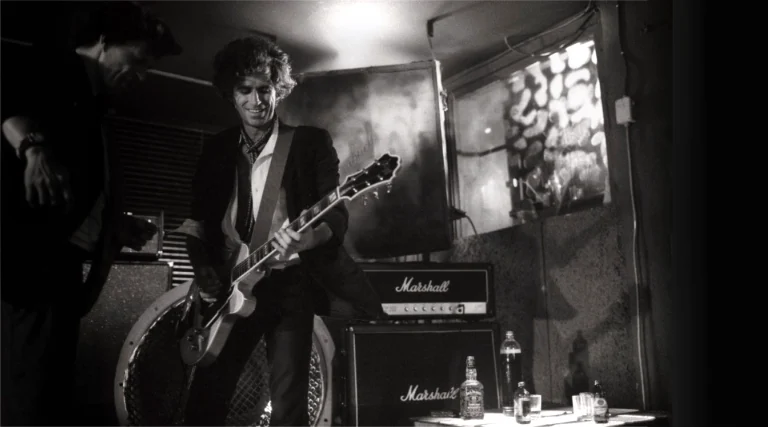 Black and white photo of a guitarist performing beside Marshall amplifiers in a smoky live music venue, capturing the nostalgic atmosphere of classic rock culture.
